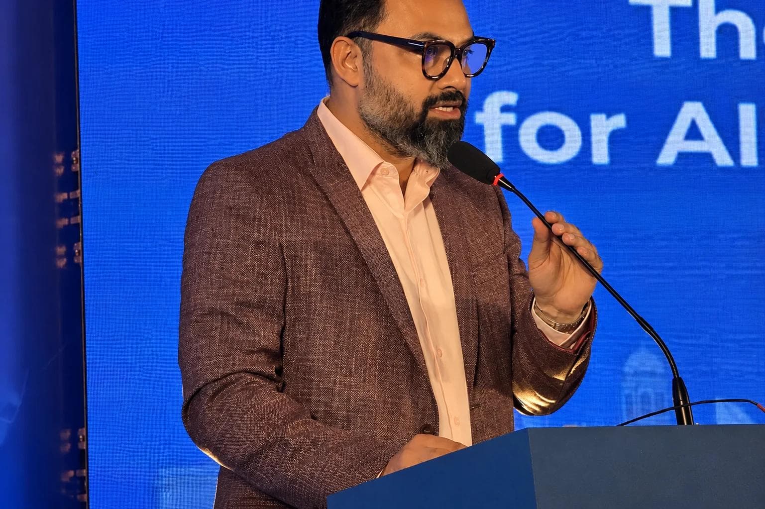 A speaker in a brown blazer at a microphone, addressing an audience with a blue presentation screen behind.