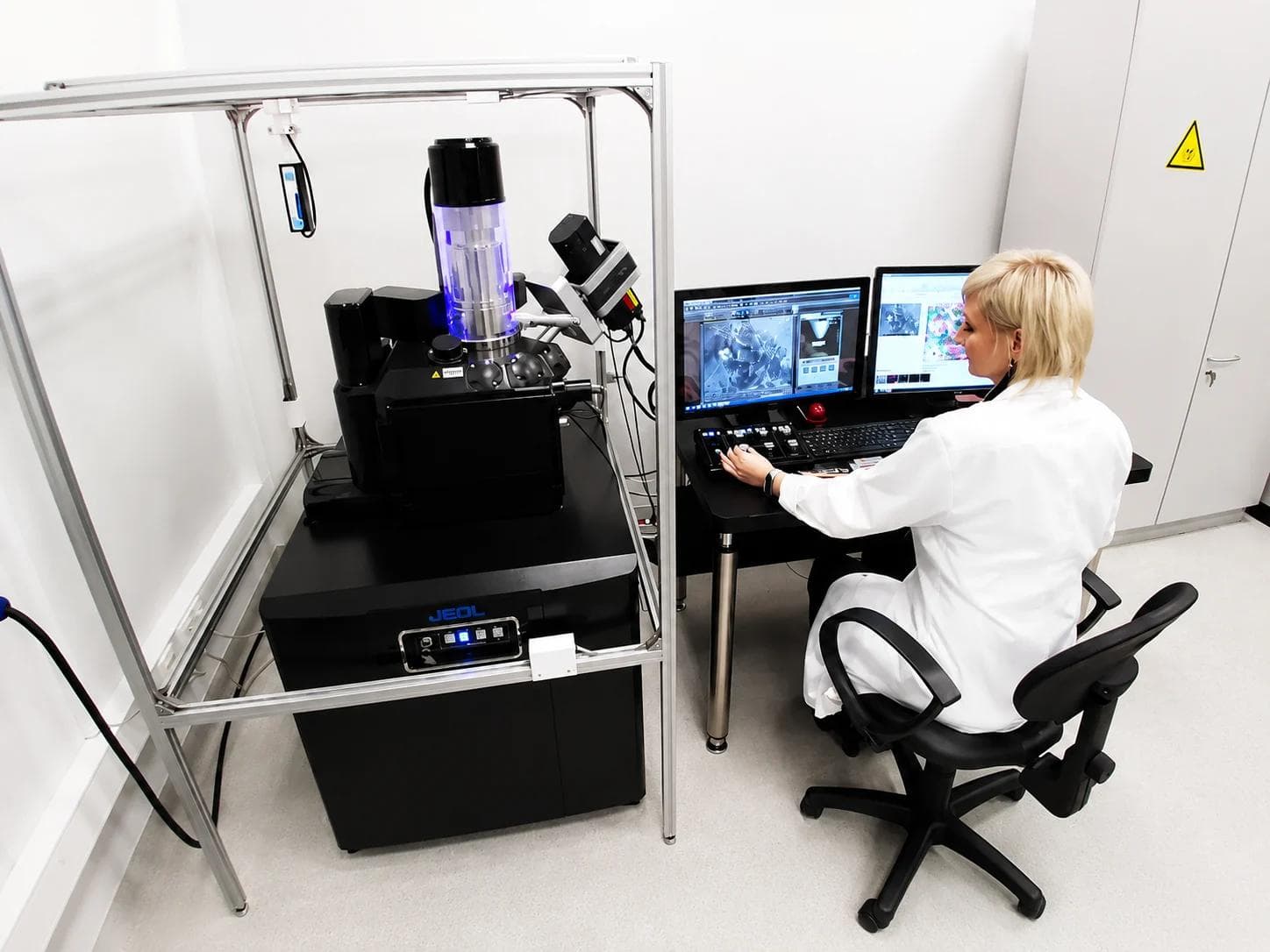 A scientist in a white coat working at the controls of a JEOL scanning electron microscope, with a monitor displaying a micrograph at the right.